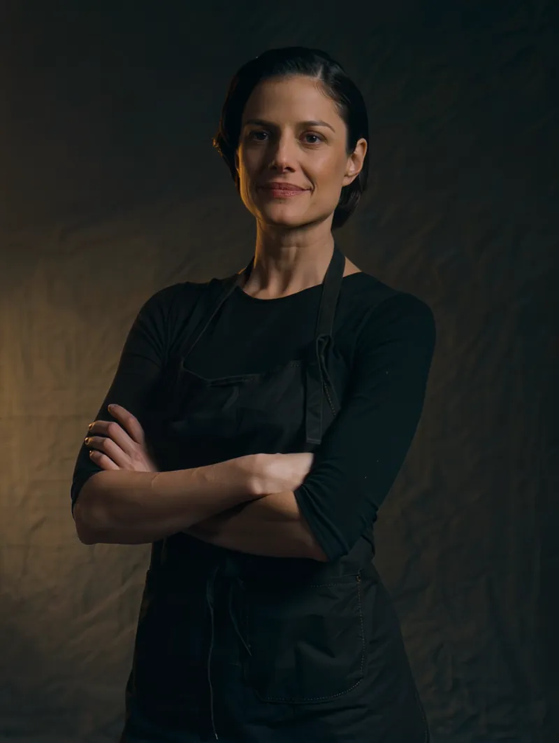 Portrait of a hair stylist in a black apron, arms crossed, lit from one side by warm brass light.